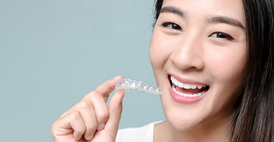 A young woman with dark hair smiling and holding a clear aligner that is part of her treatment with Invisalign in Odessa